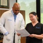 Dr. Gaetan Moise reviews a document with a colleague in an exam room, with medical scans visible on a nearby monitor.