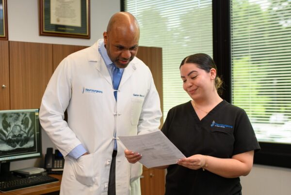 Dr. Gaetan Moise reviews a document with a colleague in an exam room, with medical scans visible on a nearby monitor.